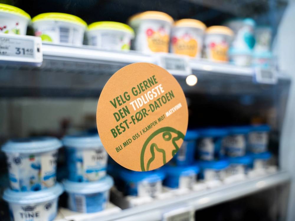 Et skilt med teksten 'Velg gjerne den tidligste best-for-datoen' henger på en kjøledisk med ulike meieriprodukter i en dagligvarebutikk.