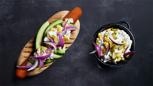 En pølse i brød med grønnsaker og løk, sammen med en skål med potetsalat på en svart bakgrunn.