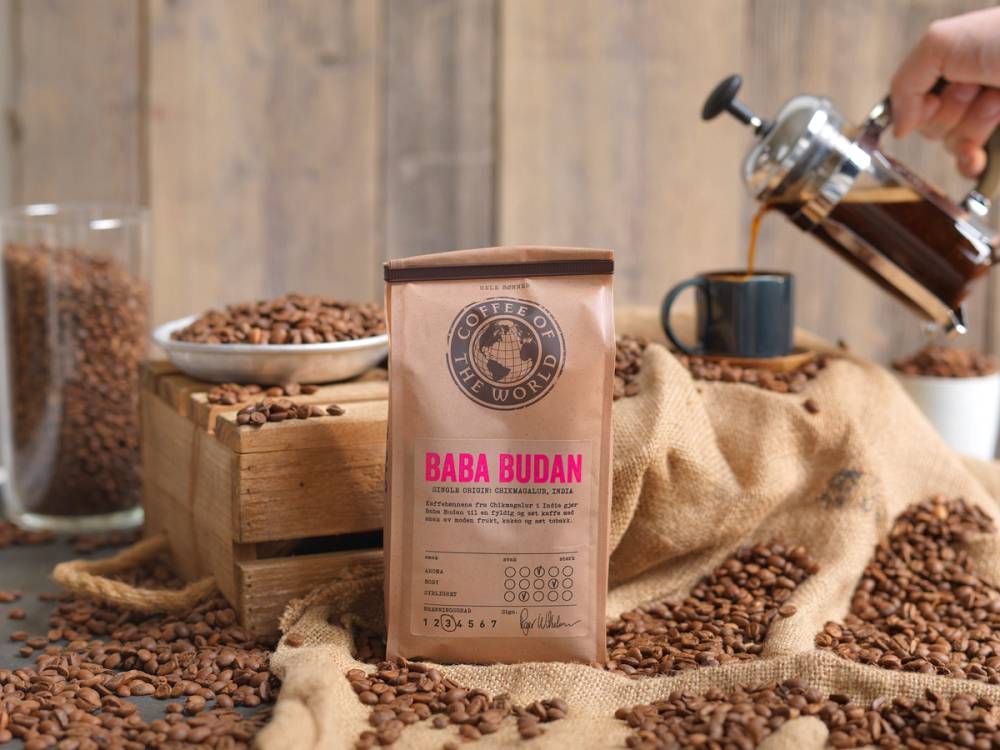 En pose med Baba Budan kaffe på et bord med kaffebønner og en kaffekopp i bakgrunnen.