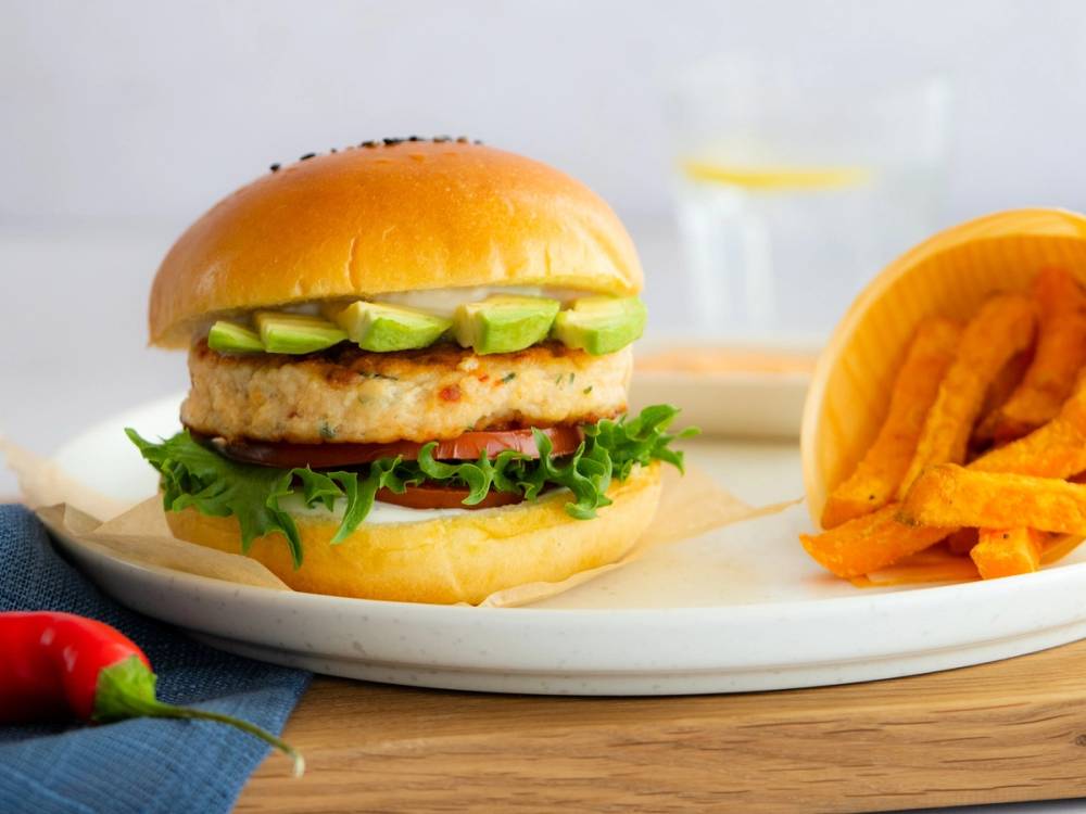 En burger med kylling, salat, agurk og en bolle, servert med pommes frites på en hvit tallerken.