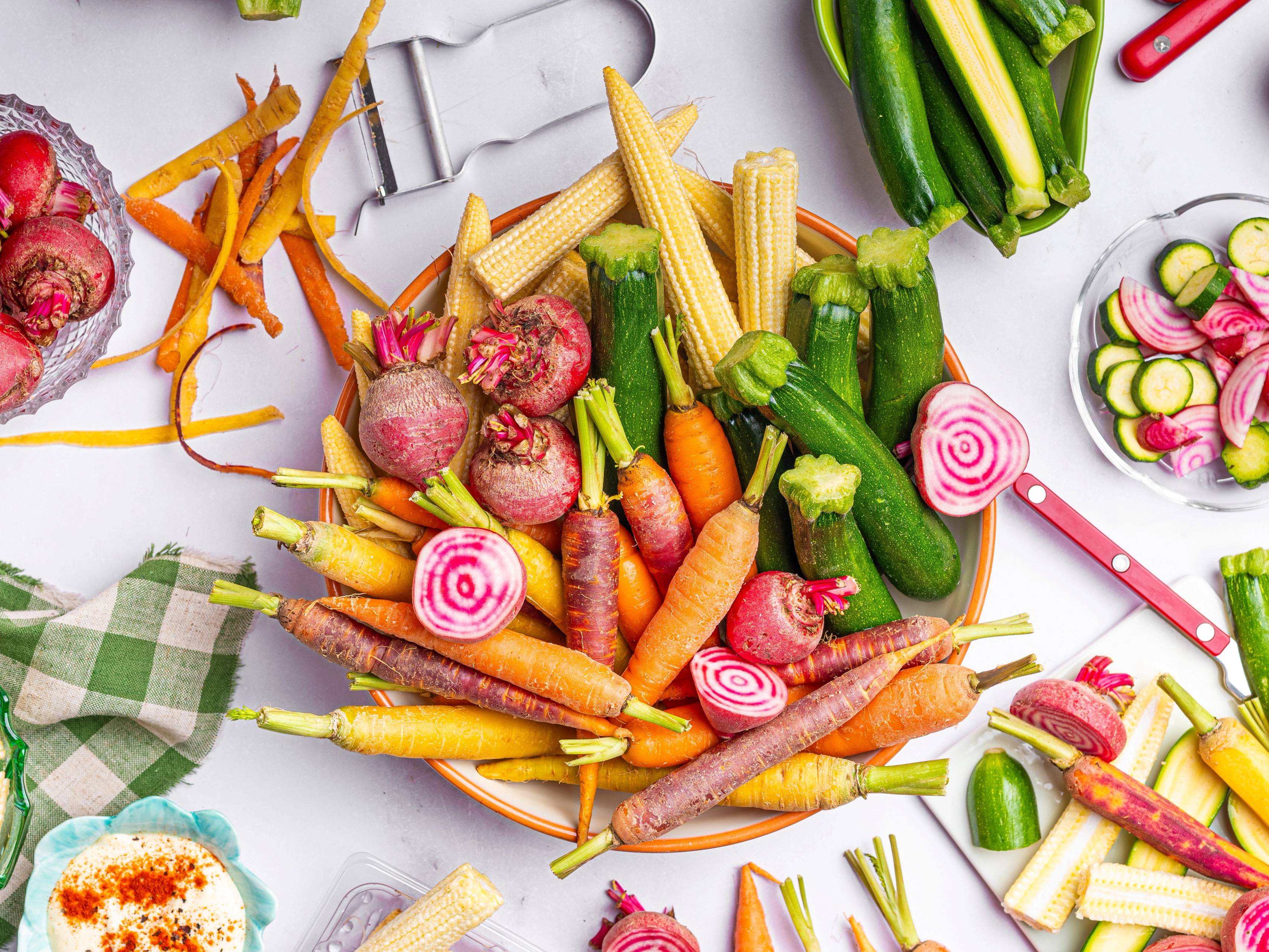 Fargerike grønnsaker som gulrøtter, reddiker og agurk ligger på en hvit overflate med en liten plakat i bakgrunnen.