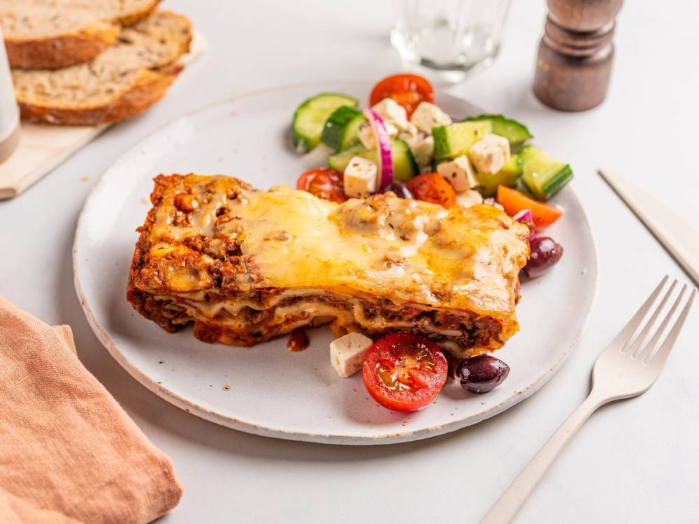 Varmeskap med lasagne og en sidesalat med agurk, tomat og oliven på en hvit tallerken