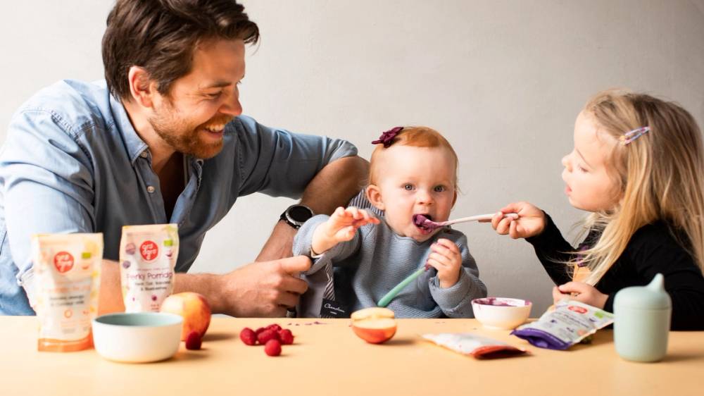 En voksen og to barn spiser sammen ved et bord med frukt og yoghurt, og barna deler mat med hverandre.