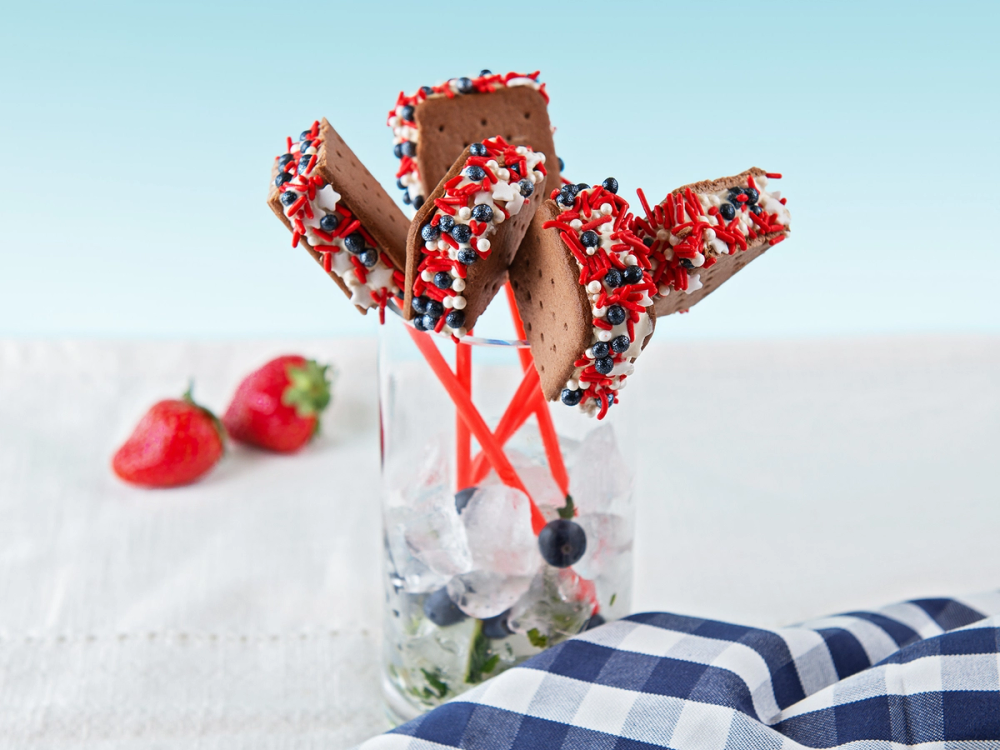 Tre iskremstenger med sjokoladetrekk og strøssel i en glass, med jordbær og blåbær ved siden av, på en hvit overflate med blått og hvitt rutete stoff i forgrunnen.