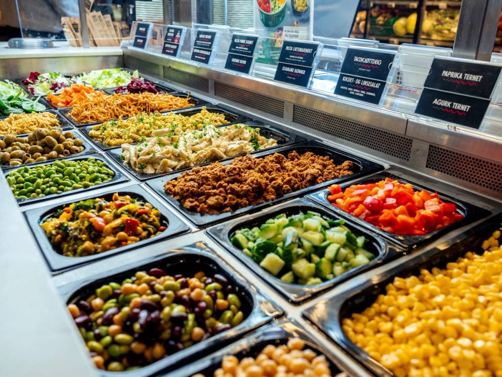 Et utvalg av ferske salater og grønnsaker i en matbutikk, med skilt over hver type.