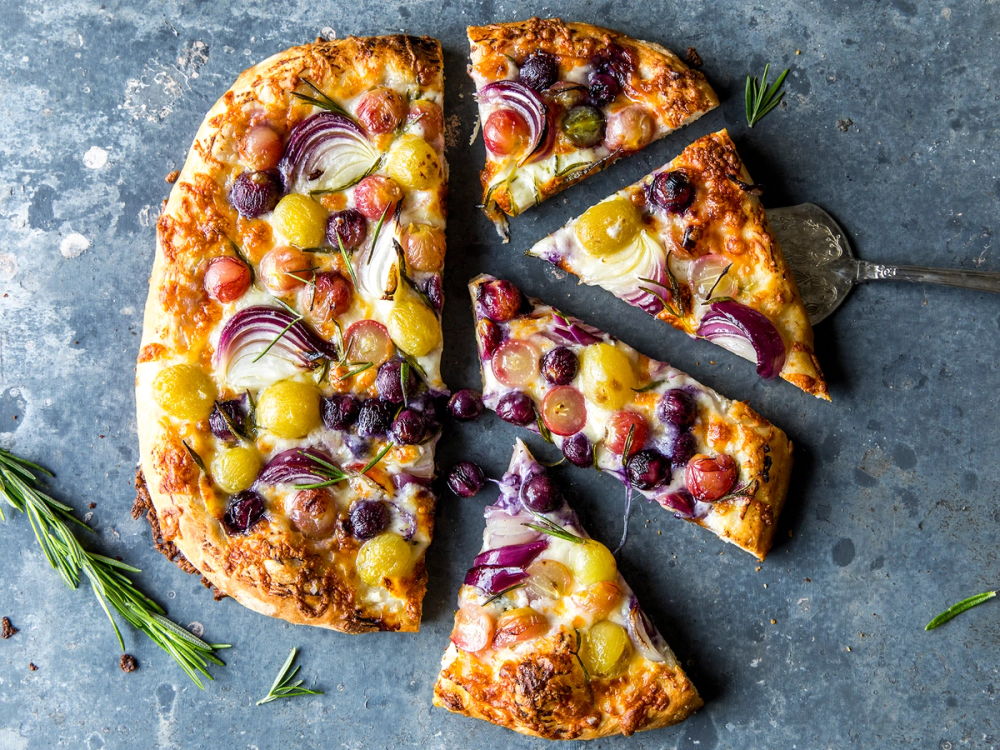 Pizza med druer, løk og ost, med en skive som er tatt ut og ligger på en pizzaspade.