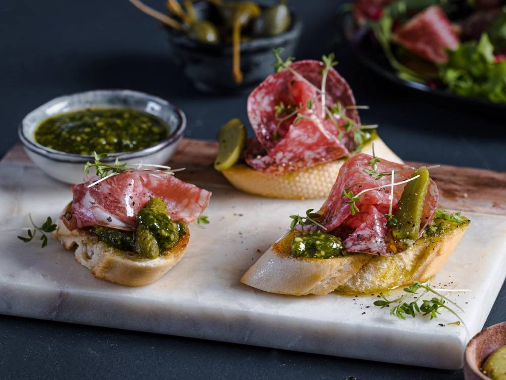 Tre bruscettaer med skinke, pesto og urter på en skjærebrett, med en liten skål med grønn saus i bakgrunnen.