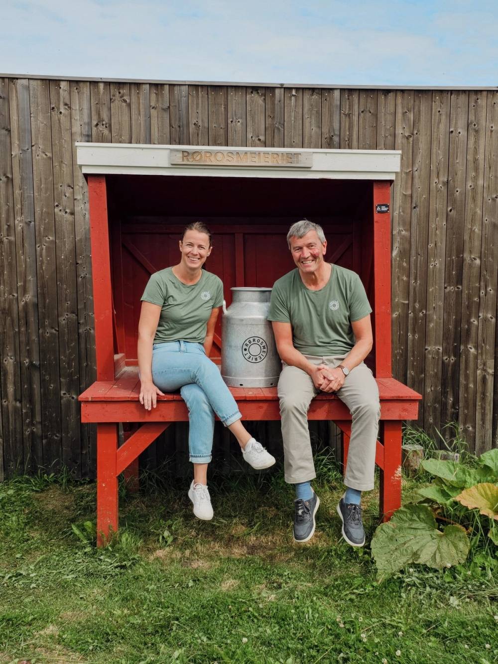 To personer sitter på en rød benk foran en trevegg, med en melkekanne mellom seg, på Rørosmeieriet.