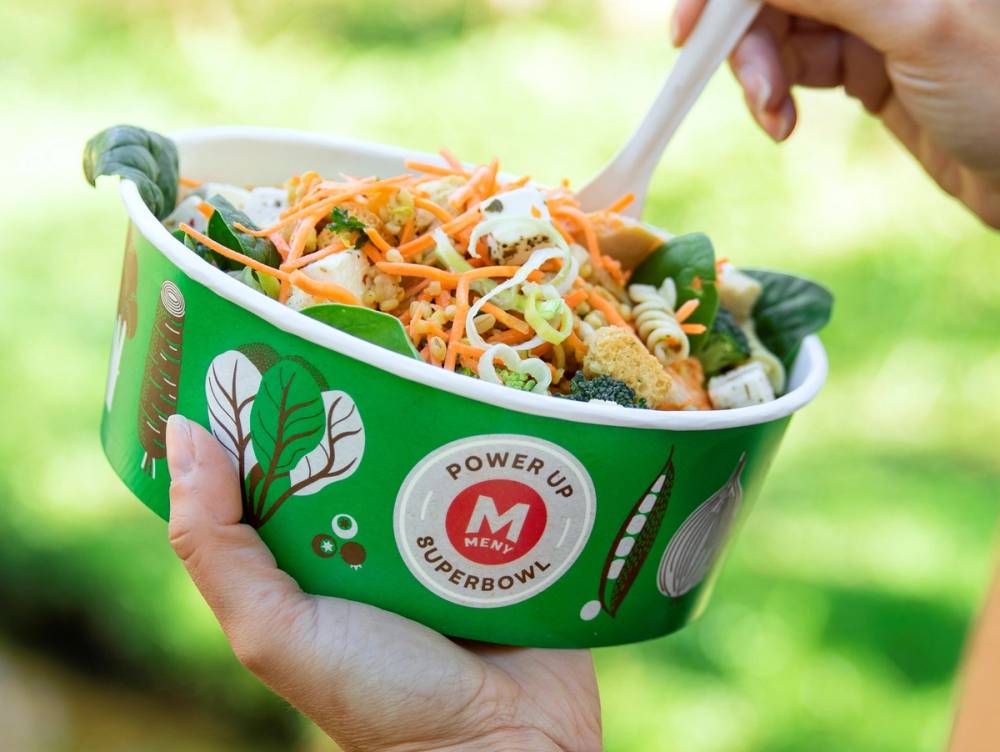 En person holder en grønn plastboks med salat og grønnsaker, og spiser med en skje utendørs.