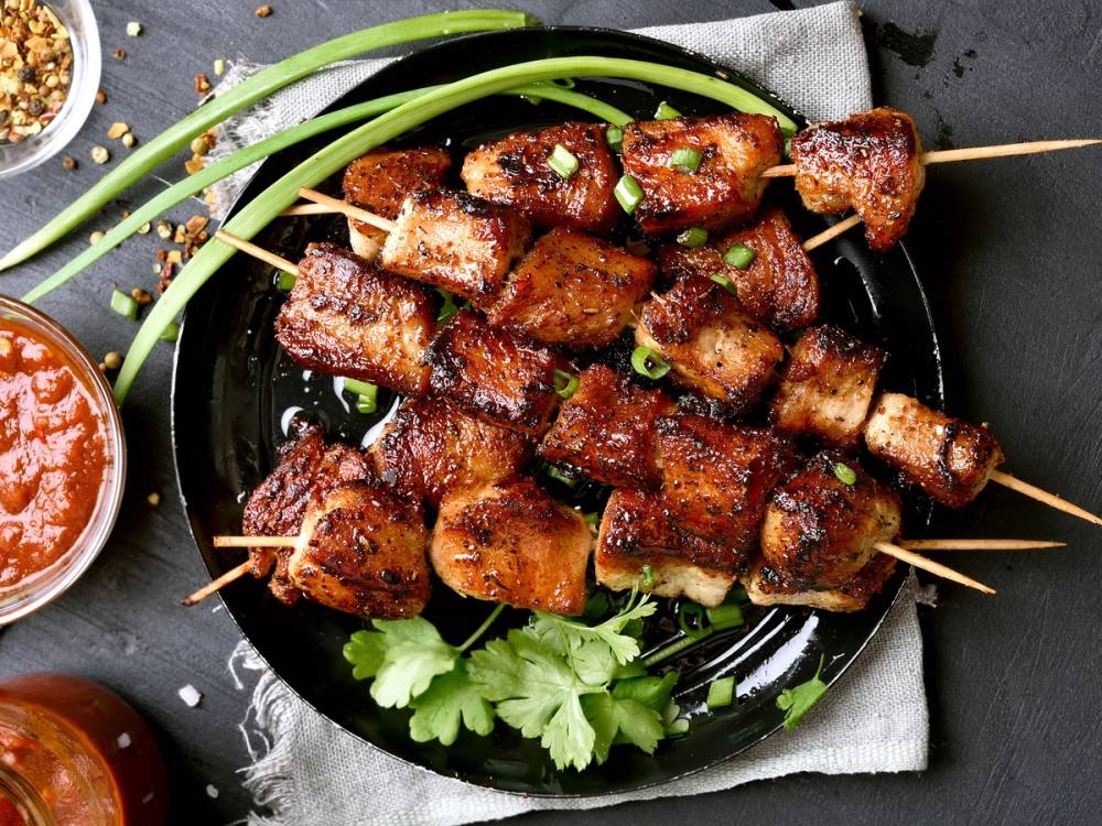 Grillede shish kebab-spyd med kjøttbiter og grønne løk på en svart tallerken, med en liten skål med saus ved siden av.