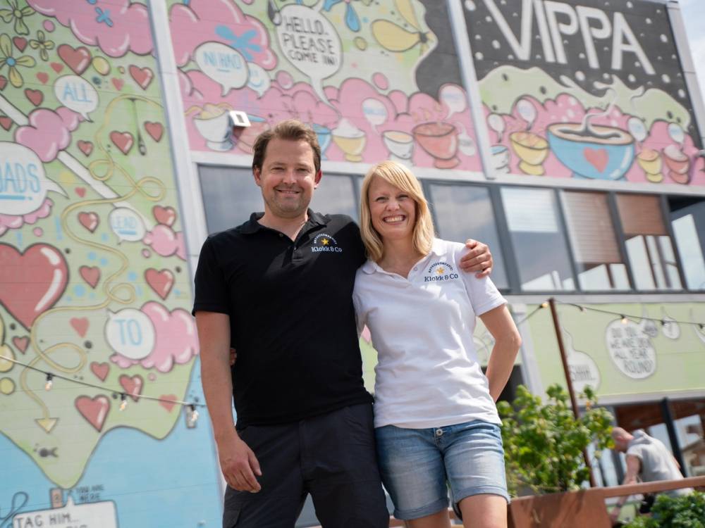 To personer står sammen foran en fargerik vegg med tegneserier og tekster, og holder rundt hverandre.