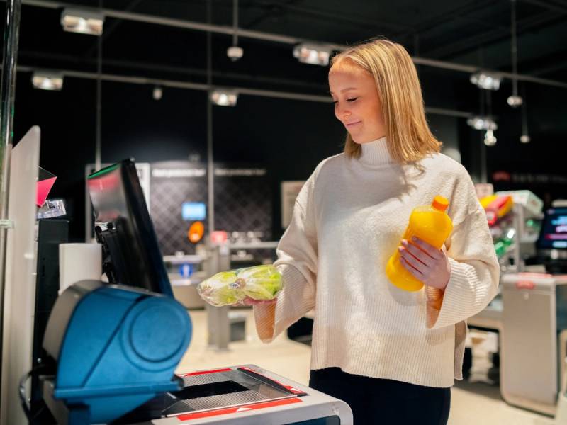 Kvinne i butikk med en flaske og en pakke i hånden, foran kassaapparat på MENY Løren.