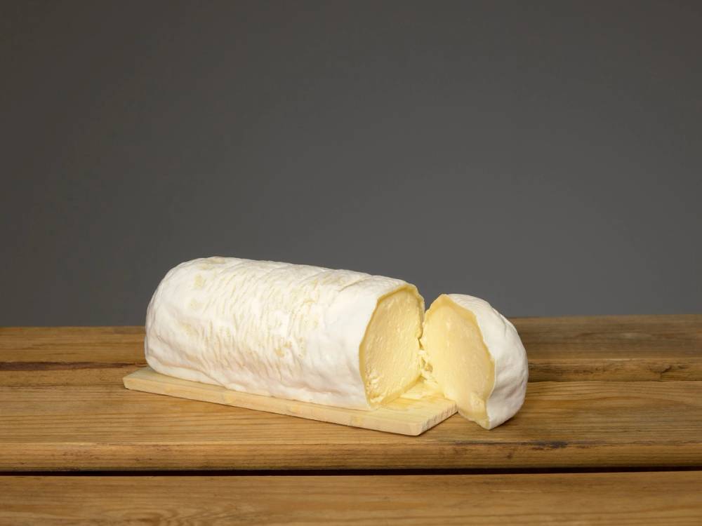 En rull med kubbeost på et trebord, med en skåret bit som viser ostens kremete innside.