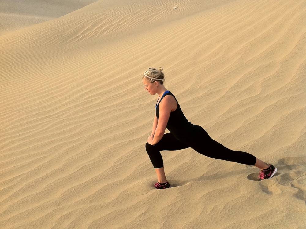 En person i svart treningstøy gjør en yoga- eller strekkøvelse på sanddyner i ørkenen.