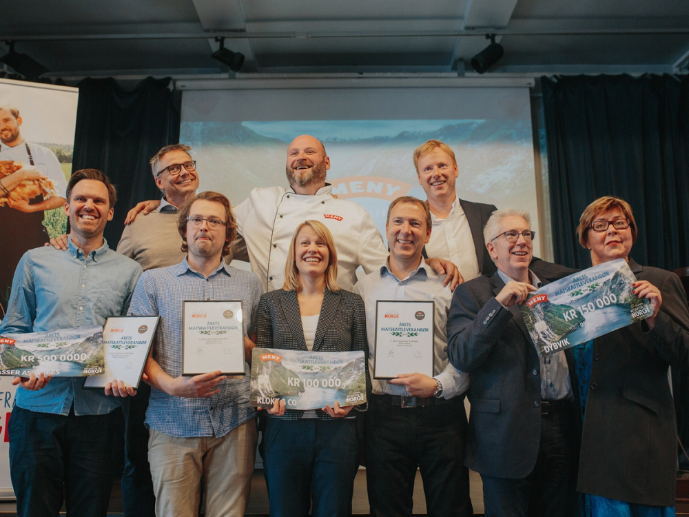 En gruppe personer på en scene som mottar priser eller diplomer under en seremoni, med en skjerm i bakgrunnen som viser en presentasjon.