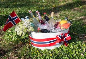 En hvit bøtte med norske flagg og blomster, fylt med is og ulike drikkevarer, plassert på gresset til 17. mai-feiring.