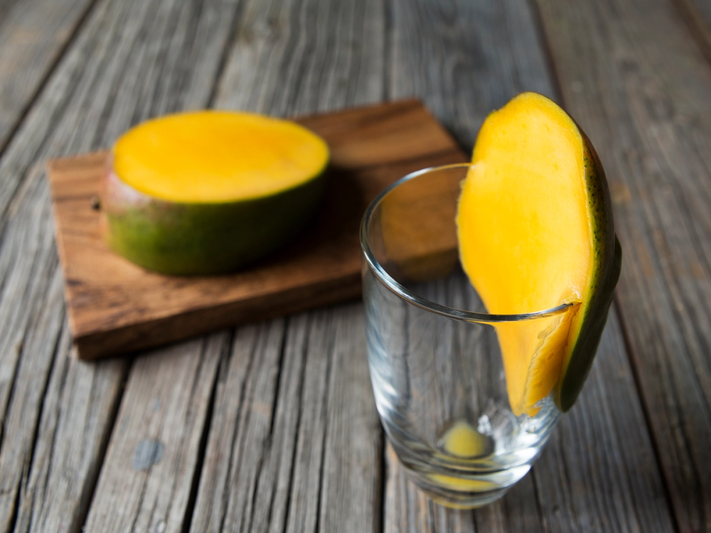 En skrelt mango skåret i en skive som står i et glass, med en halvmåneformet mango på en skjærefjøl i bakgrunnen.