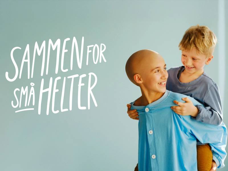To barn, ett med raket på klærne, omfavner hverandre foran en lys blå vegg med teksten 'Sammen for små helter'.