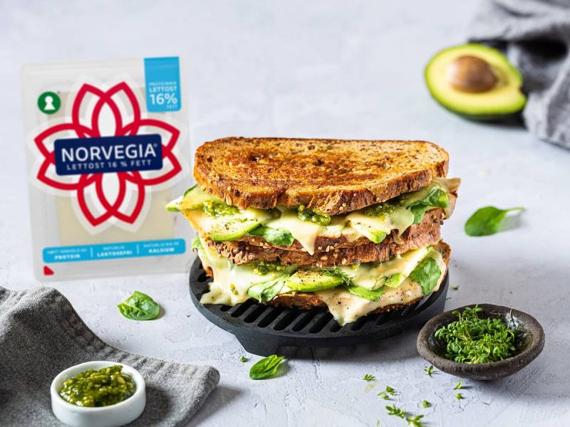 En toast med avokado og salat på en svart grillpanne, omgitt av avokadobiter og små skåler med grønn saus, med en melkekartong i bakgrunnen.