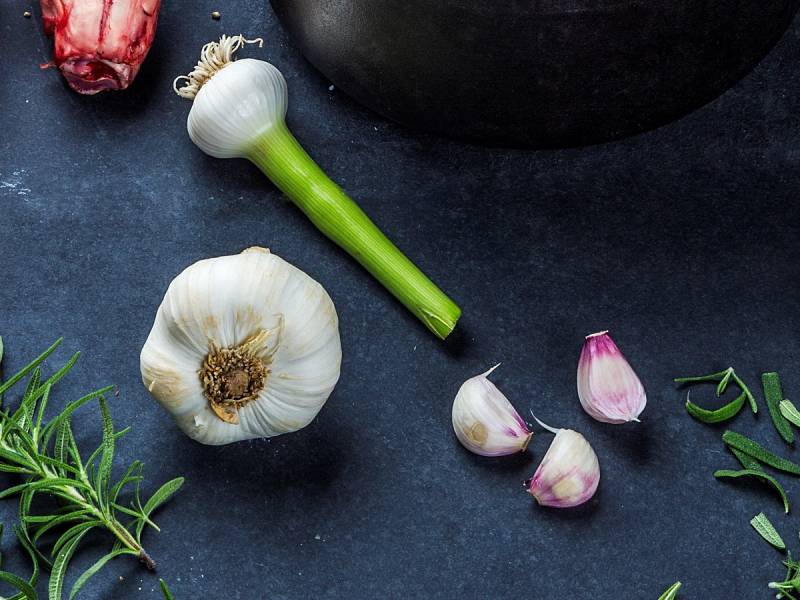 Hvitløk, hvitløkfedd og frisk rosmarin ligger på en mørk overflate, med en svart gryte i bakgrunnen.
