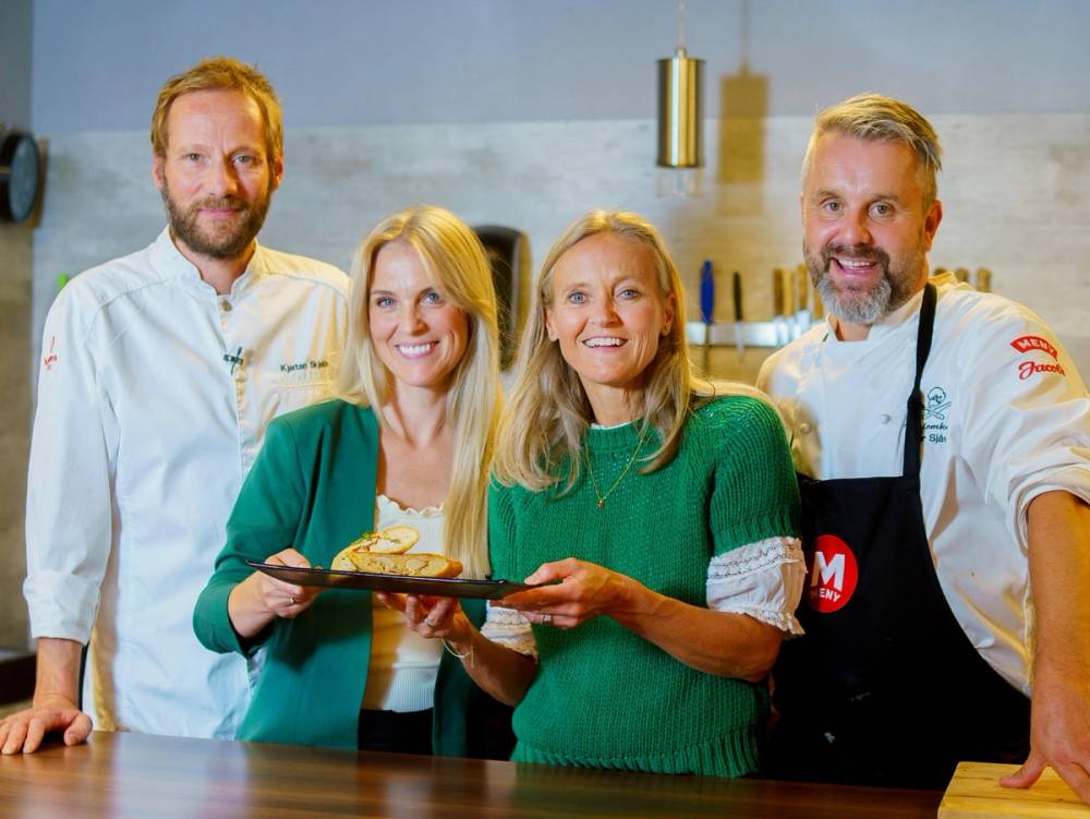 To personer holder en tallerken med mat mens de står i et kjøkkenmiljø, med en kokk i bakgrunnen.