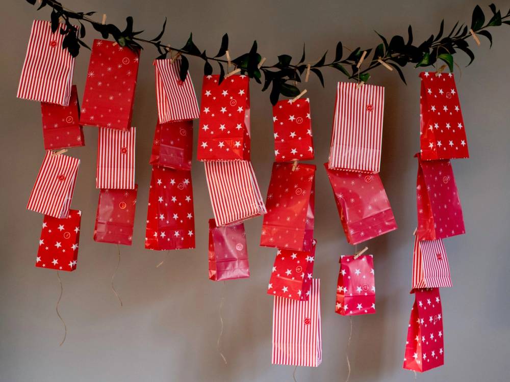 Julepynt med små gaveposer og striper i rødt og hvitt henger fra en grein mot en vegg.