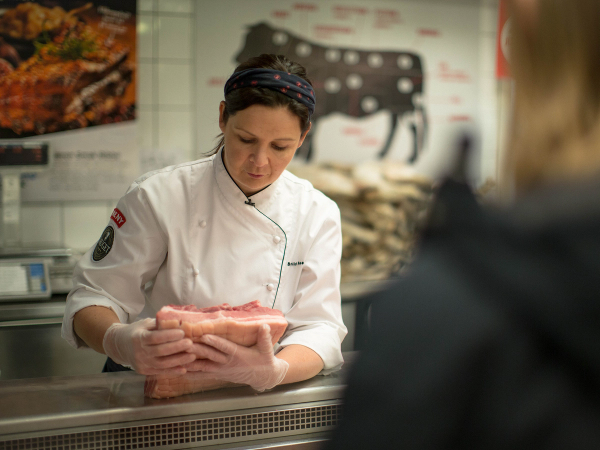 Kokk i hvit uniform og svart lue som viser fram en pakke med kjøtt til en kunde i en matbutikk eller på markedet, med en meny eller reklameplakat i bakgrunnen.
