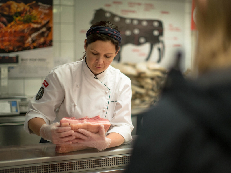 Kokk i hvit uniform og svart lue som viser fram en pakke med kjøtt til en kunde i en matbutikk eller på markedet, med en meny eller reklameplakat i bakgrunnen.