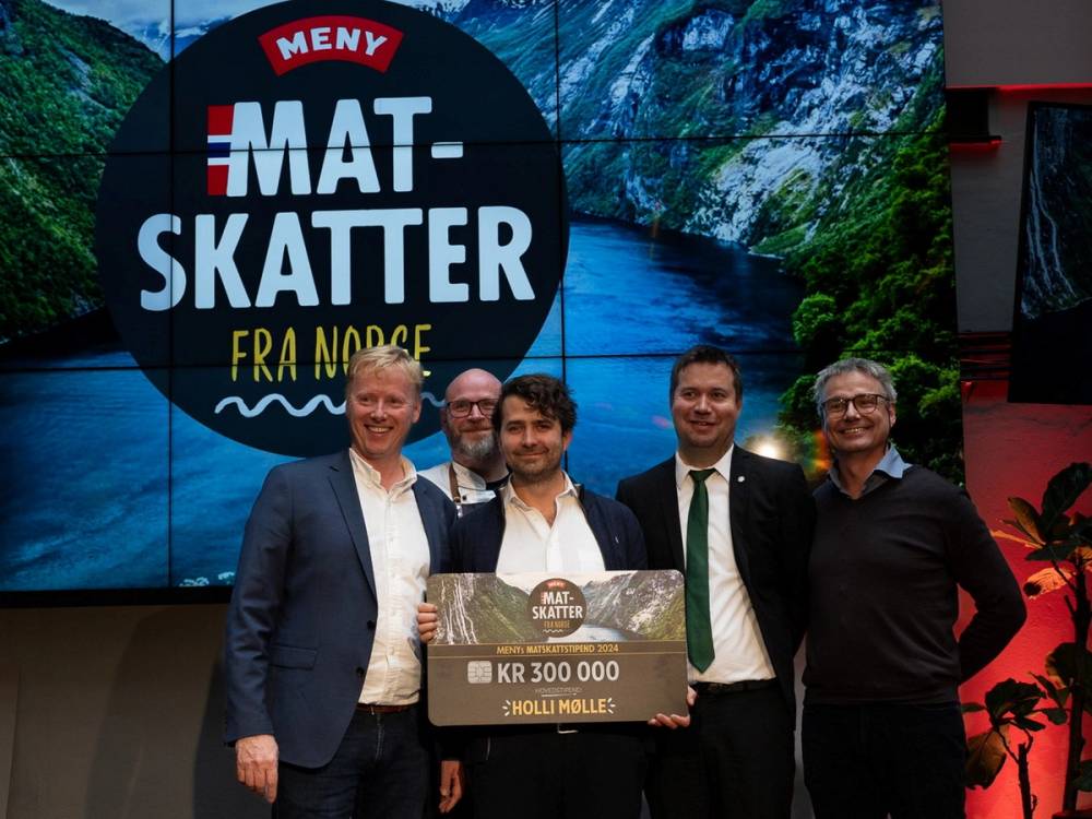 Fire personer står på scenen med en stor sjekk og en banner som sier 'Mat- Skatter fra Nord', med fjell og elv i bakgrunnen.