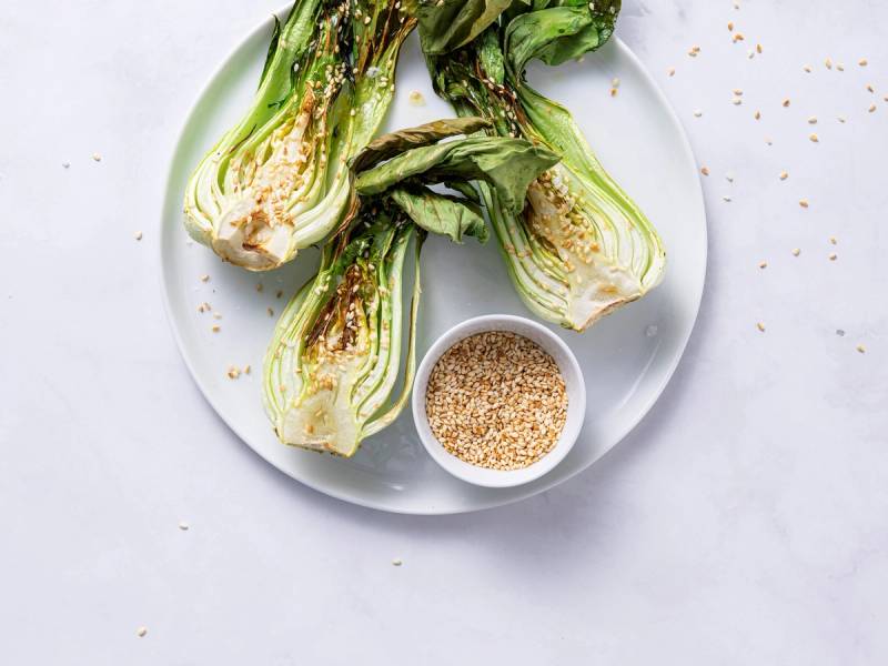 Pak choy med sesamolje på en hvit tallerken, med en liten skål med sesamfrø ved siden av.