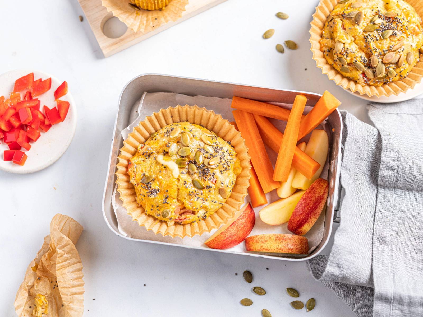 Muffins i papirformer med gul ost og frø, servert med gulrotstaver og epleskiver i en matboks, omgitt av små gresskarkjerner og en liten skål med hakket tomat.