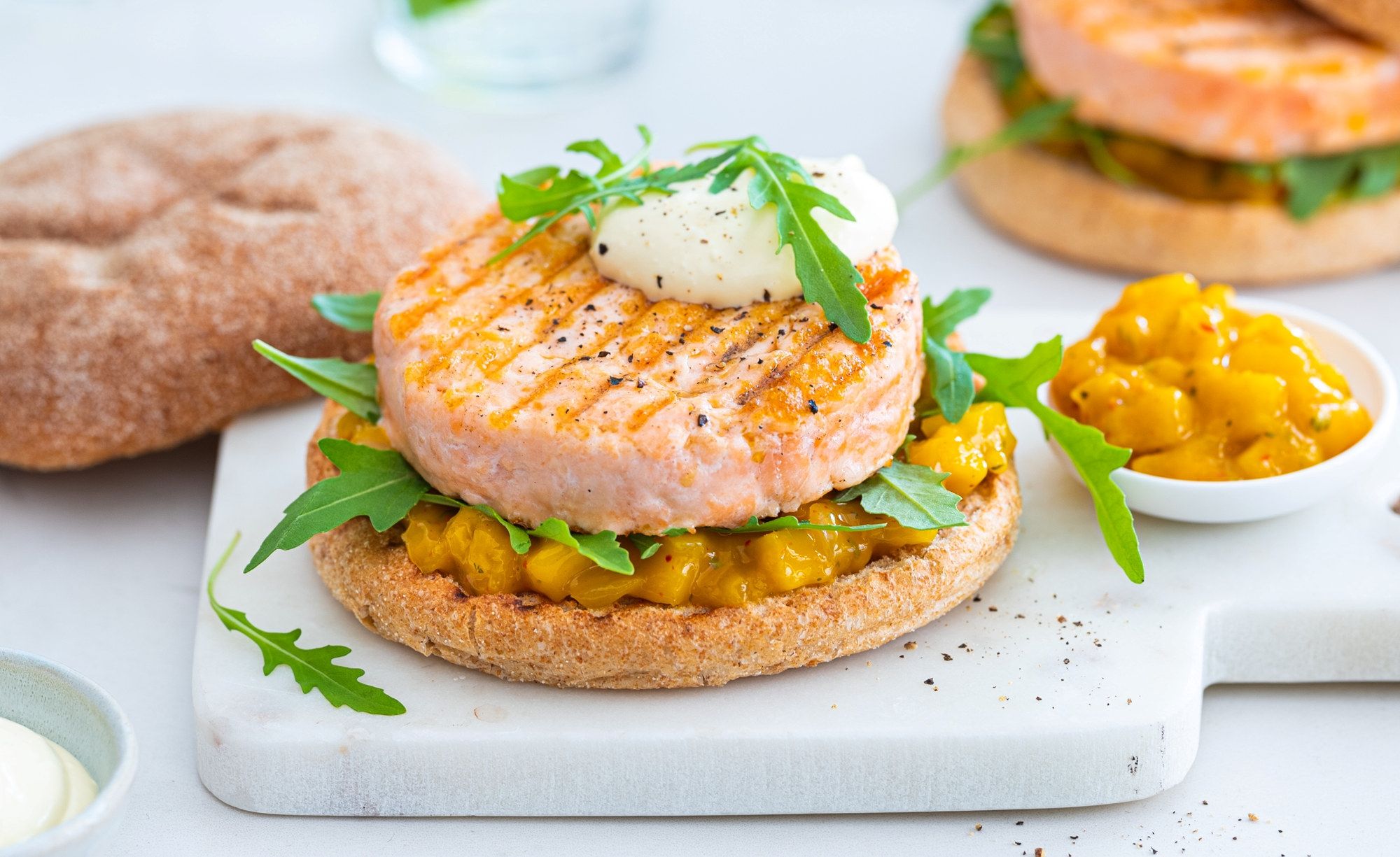 Lakseburger med mango, toppet med en klatt rømme og frisk grønn urter, servert på en flat brødskive med en side av mango- og sitrusdressing.