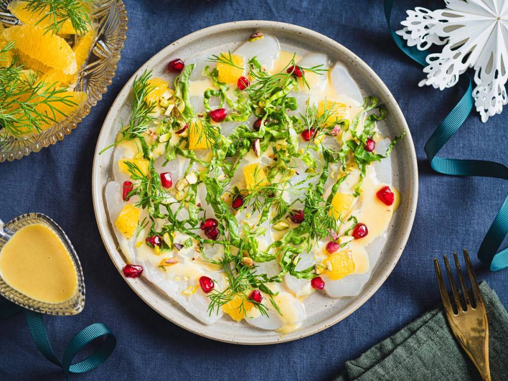 En tallerken med kveite marinert med appelsin, pyntet med grønt og granateplefrø, omgitt av sitronbiter og en liten saus eller dressing på siden.