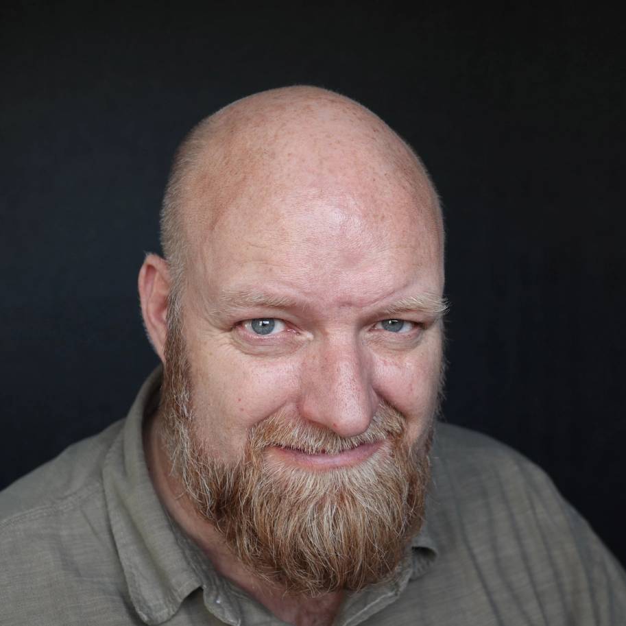 En mann med skjegg og lys genser, sett forfra mot en mørk bakgrunn.