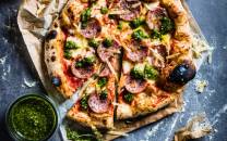 Pizza med skinke, brokkoli og ost på en treplate, med en liten skål med grønn saus ved siden av.