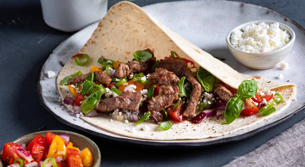 Strimler av lammelår i en tortilla med grønnsaker og en liten skål med hvit saus ved siden av.