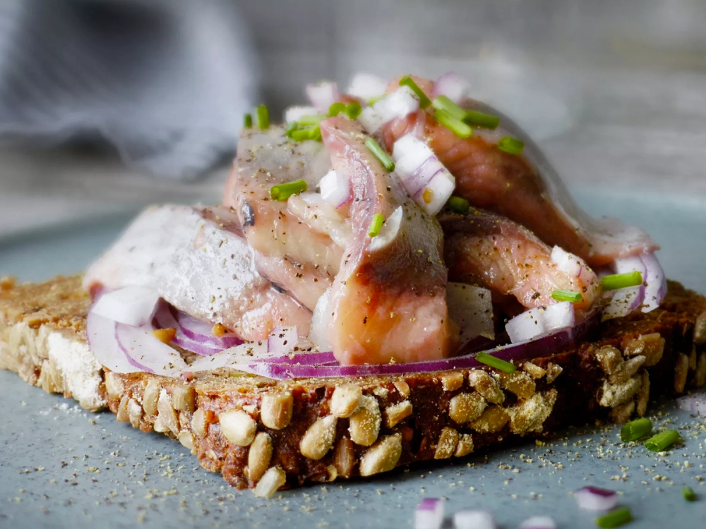 En åpen sandwich med sild, rødløk og grønne urter på en brødskive med frø og nøtter, klar til servering.
