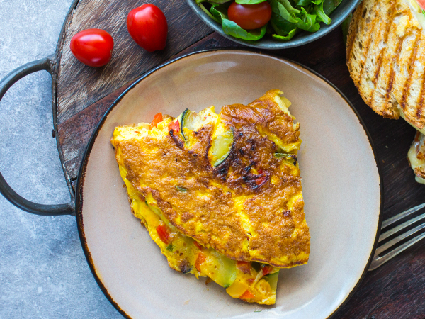 En omelett med paprika og squash på en tallerken, med tomater og brød i bakgrunnen.