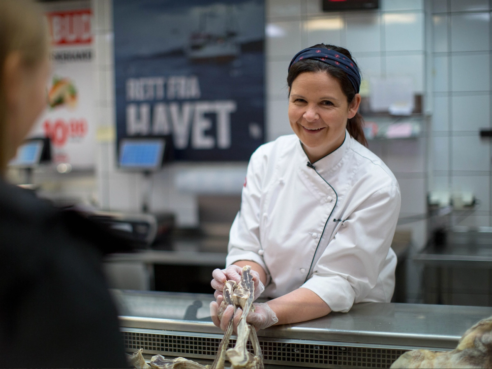 En kokk i hvit uniform og blå forkle som håndterer sjømat i en butikk eller restaurantkjøkken, med en kunde i forgrunnen.