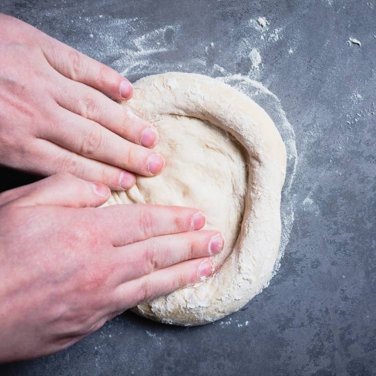 Hender former en rund pizzadeig på en melet arbeidsflate.