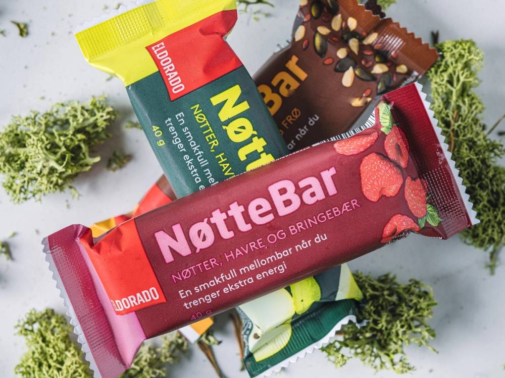 Flere NotteBar energibarer i ulike smaker ligger på en hvit overflate med grønt dekorativt blad rundt.