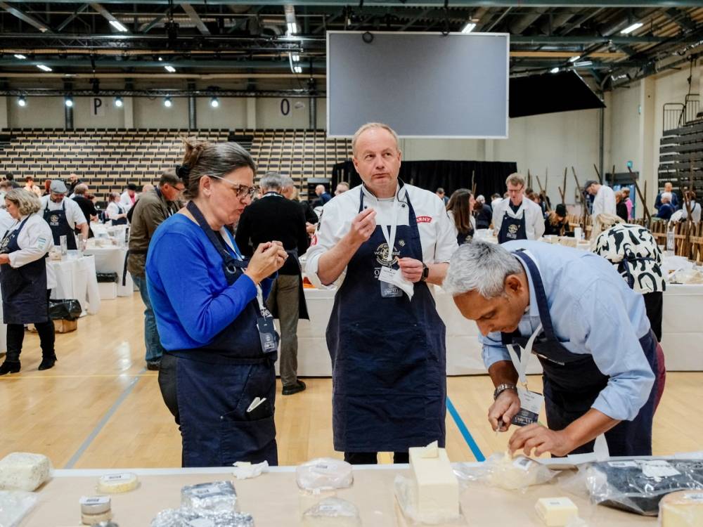 Personer som deltar på en ostemesse, med fokus på en person som skjærer ost, i et stort innendørs arrangement.