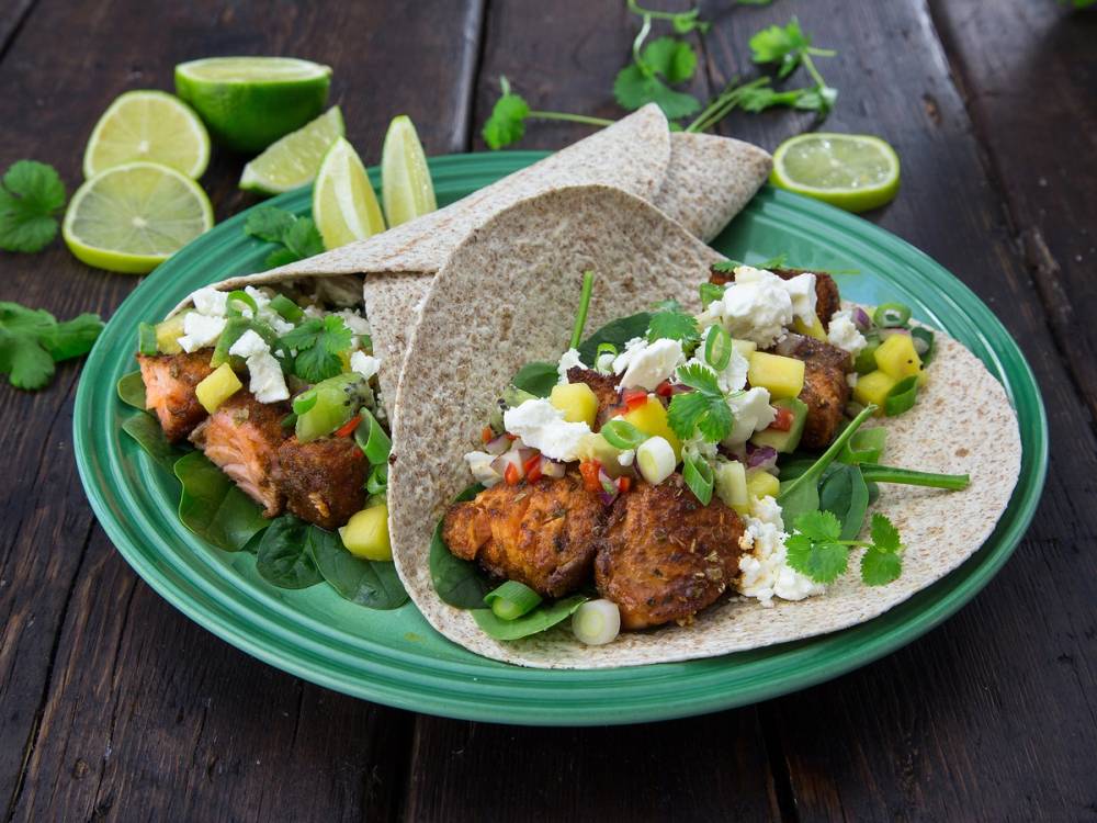 Fisketacos med grønnsaker, ost og lime på en grønn tallerken, omgitt av limebåter og urter.