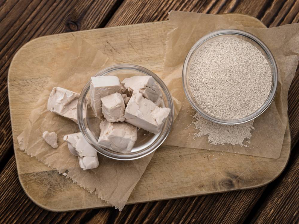 Tørrgjær og fersk gjær i glassboller på et trebord, med tørrgjær i en skål og fersk gjær i en annen, begge på papirark.