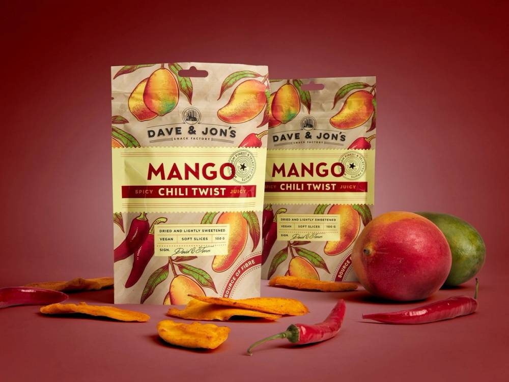 To pakker med mango-chili twist snacks, to mangoer og en chilipepper på en bordplate mot en rød bakgrunn.