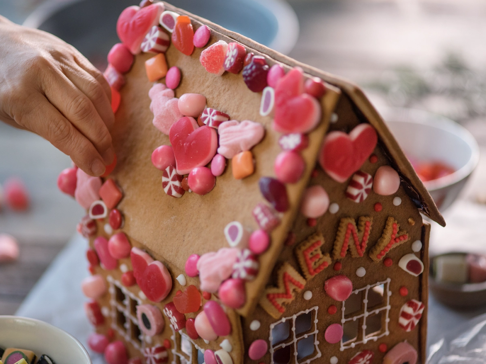 En person legger til pynt på et pepperkakehus dekorert med ulike rosa og røde godterier og små sukkerskruer.