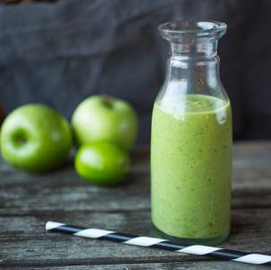 En grønn smoothie i en glassflaske med to grønne epler ved siden av på et treverkebord.