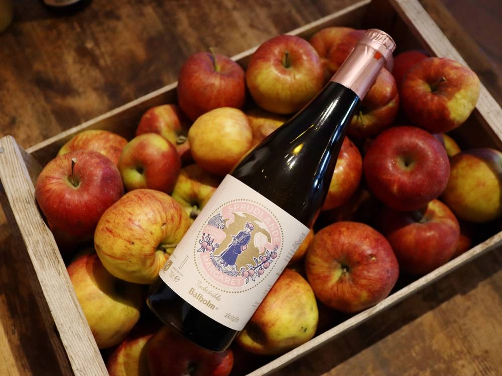 En eske med mange røde og gule epler og en flaske med vin oppi, plassert på en treflate.