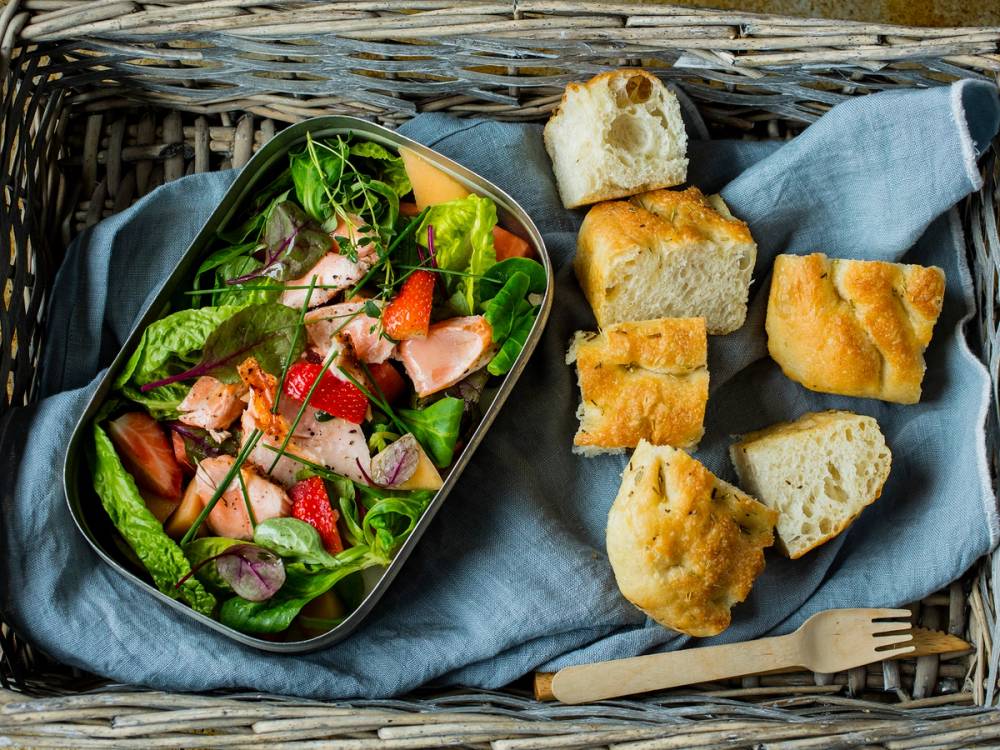 En salat med grønne blader, jordbær og skinke, sammen med flere stykker focaccia på en blå duk i et kurvaktig underlag.