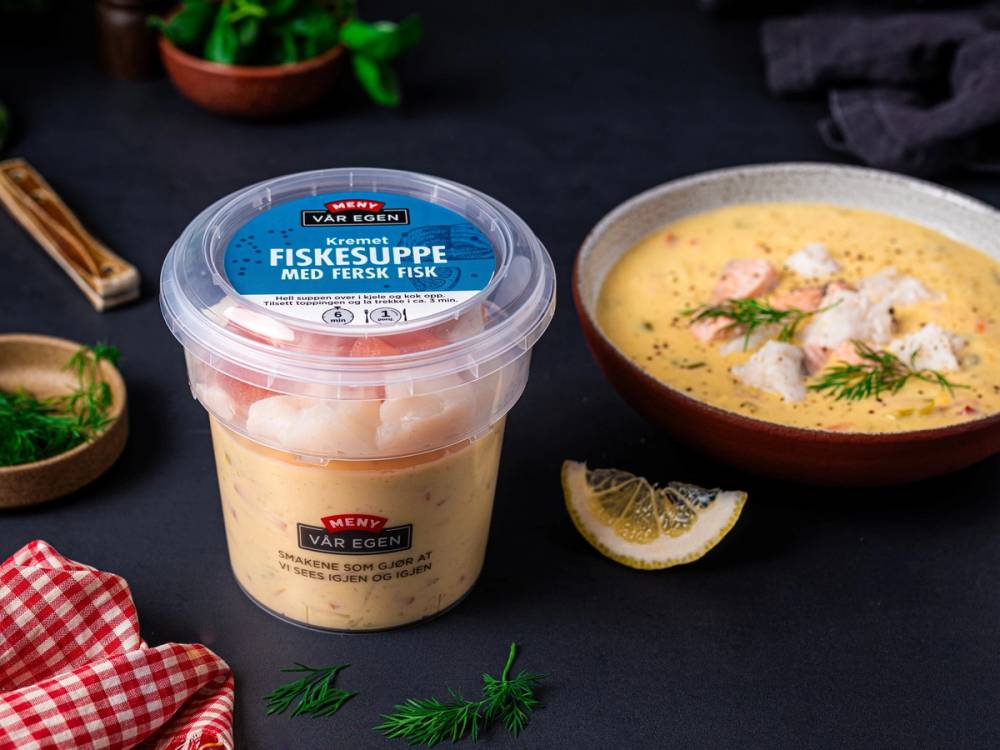 Fiskesuppe med fersk fisk servert i en bolle med sitronbåt ved siden av, omgitt av friske urter og en plastbeholder med fiskesuppe